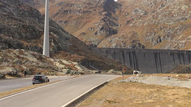 Red car driving on a mountain pass road at Gotthard Pass, Switzerland. Static shot of traffic passing a concrete dam wall in the Swiss Alps, autumn slopes and alpine road