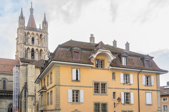 View of the historic Lausanne Cathedral rising above a traditional yellow building with terracotta roof tiles and white shutters in Lausanne, Vaud, Switzerland.