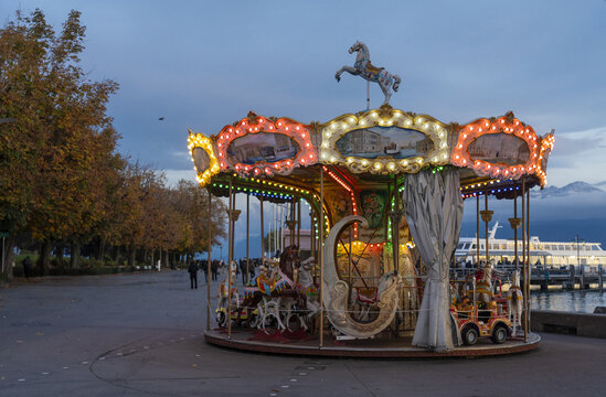 View of an illuminated carousel by the lake with autumn trees and mountains in the background in Lausanne, Vaud, Switzerland.