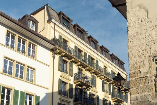 View of historic residential buildings with yellow and white facades, green shutters, and wrought iron balconies under a clear blue sky in Lausanne, Vaud, Switzerland.