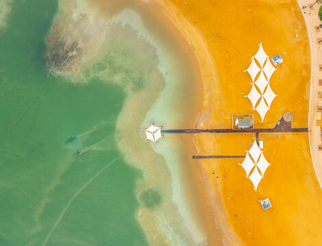 Aerial view of the Dead Sea shoreline with white sunshades on a sandy beach and a wooden pier extending into the turquoise water in Beersheba, South District, Israel.