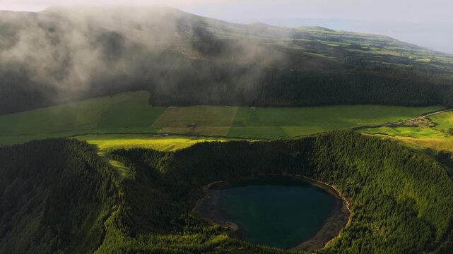 Aerial view of the volcanic crater lake in Sete Cidades surrounded by lush green forests and rolling hills under a blanket of low clouds in Sete Cidades, Azores, Portugal.