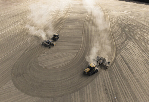Aerial view of two tractors leveling a large, dusty agricultural field, creating curved patterns in the soil in Wieringerwaard, North Holland, Netherlands.