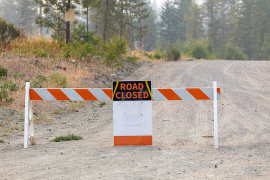 A road closed sign and barricade block a gravel forest road due to wildfire hazard and smoky haze in Kelowna, British Columbia.Road closed sign blocking a dirt road due to wildfire danger and smoke