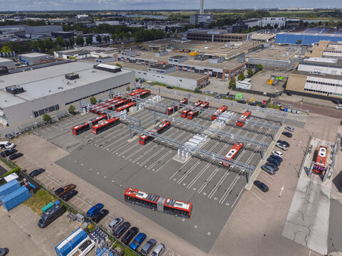 Aerial view of a bus depot with red electric buses parked in bays with charging infrastructure, surrounded by industrial buildings in Haarlem, Noord-Holland, Netherlands.