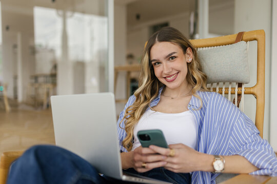 Smiling woman works on laptop and texts on smartphone at home