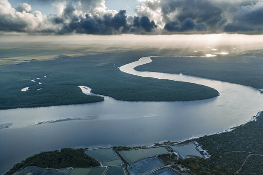 Aerial view of Pontal da Barra do Mamanguape featuring the winding Mamanguape River through lush mangroves under dramatic clouds with sunbeams in Rio Tinto, State of Paraiba, Brazil.
