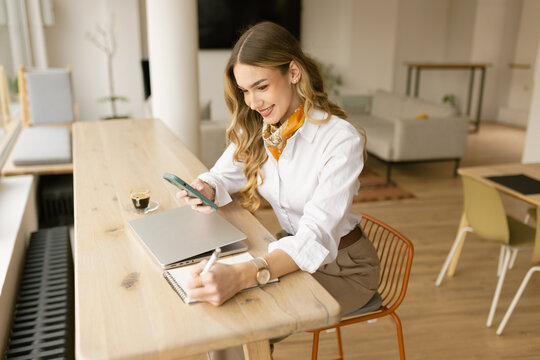 Smiling female entrepreneur texting and taking notes in modern office
