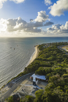 Aerial view of Ponta do Seixas lighthouse surrounded by lush coastal vegetation, a winding road, and the Atlantic Ocean under a bright sky in Joao Pessoa, State of Paraiba, Brazil.