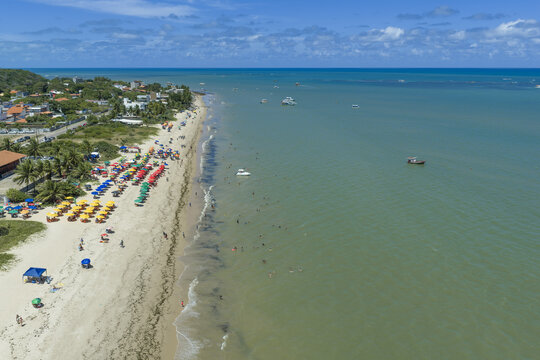 Aerial view of Seixas Beach with colorful umbrellas lining the sandy shore, people swimming in the turquoise water, and small boats anchored offshore in Joao Pessoa, Paraiba, Brazil.