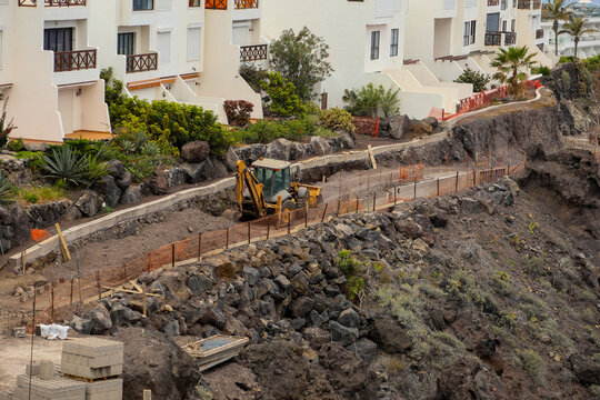 View of a yellow backhoe loader working on a construction site along a rocky volcanic cliffside with white residential buildings in Santa Cruz de Tenerife, Canary Islands, Spain.