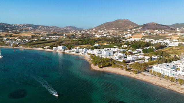Aerial view of the coastal town of Paros with white buildings, sandy beach, turquoise waters, and a small boat cruising along the shoreline in Paros, Greece.