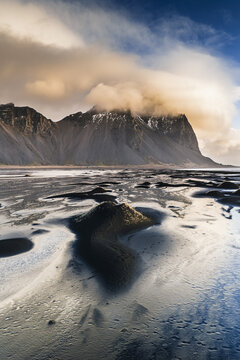 Aerial view of Vestrahorn mountain rising above the dramatic black sand beach with textured dunes and soft golden light under a cloudy sky, Sveitarfelagid Hornafjordur, Iceland.