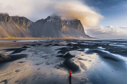 Aerial view of Vestrahorn mountain rising above the black sand beach with a person in an orange jacket walking near tidal pools in Sveitarfelagid Hornafjordur, Iceland.