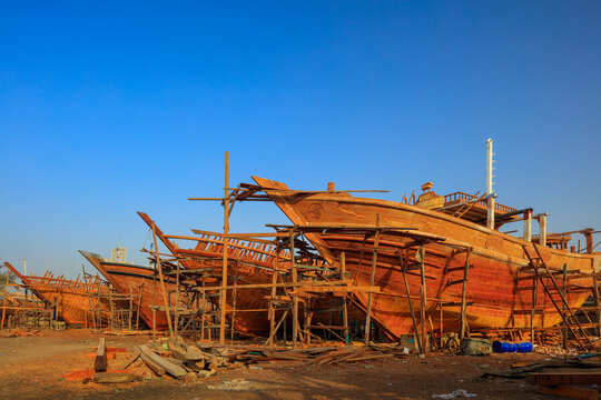 View of traditional wooden dhows under construction in a shipyard, supported by wooden scaffolding under a clear blue sky in Karachi, Sindh, Pakistan.