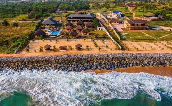 Aerial view of a coastal resort in Ghana featuring thatched-roof bungalows, a swimming pool, and a stone sea wall along the sandy beach.