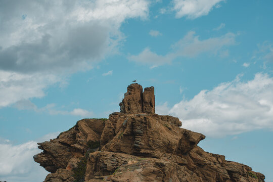 View of the rocky cliffs of Cala des Murtar with a bird perched on the summit under a cloudy blue sky, Illes Balears, Islas Baleares, Spain.
