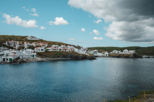 View of the coastal village of Cala des Murtar with white houses nestled on rocky cliffs overlooking the blue sea under a cloudy sky in Illes Balears, Islas Baleares, Spain.
