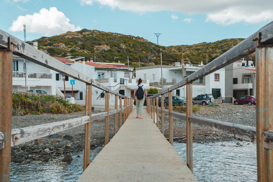 View of a woman walking along a concrete pier towards the white houses of the coastal village of Cala des Murtar, Illes Balears, Islas Baleares, Spain.