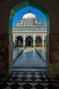 View of the ornate Shrine of Shah Abdul Latif Bhittai framed by a stone archway with intricate blue tile work and a white dome in the marble courtyard, Bhit Shah, Sindh, Pakistan.