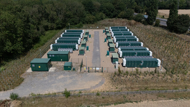 Aerial view of a data centre facility with rows of green storage containers on a gravel lot, enclosed by a security fence and surrounded by trees in Hatfield, England, United Kingdom.