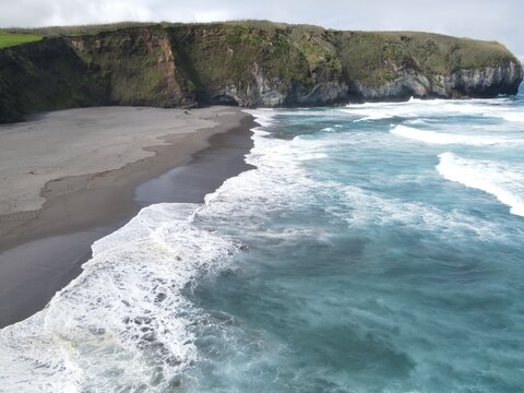 Aerial view of a black sand beach nestled against dramatic cliffs with crashing Atlantic ocean waves in Azores, Portugal.