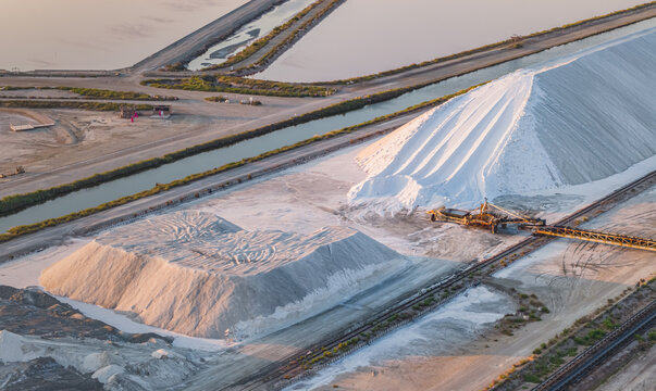 Aerial view of massive sea salt stockpiles connected by conveyor systems at an industrial salt production facility during golden hour in France.
