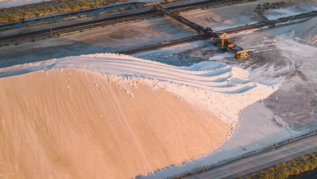 Aerial view of a massive sea salt stockpile illuminated by golden hour light at an industrial salt production facility, featuring a textured mineral mound and conveyor belt system in France.