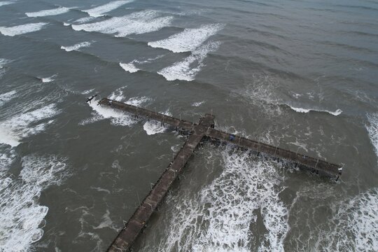 Aerial view of the T-shaped pier extending into the Atlantic Ocean with waves crashing against the concrete structure in Mongagua, State of Sao Paulo, Brazil.