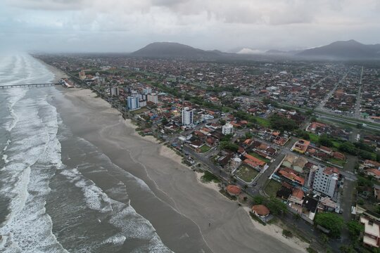 Aerial view of the coastal town of Mongagua, featuring the sandy beach, ocean waves, residential buildings, and distant mountains under a cloudy sky in Mongagua, State of Sao Paulo, Brazil.