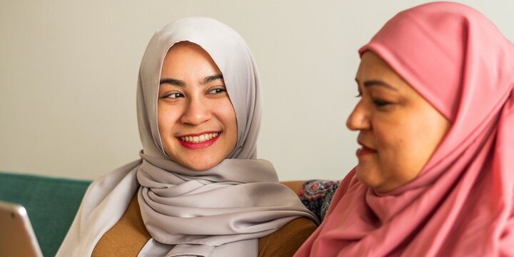 Two women in hijabs smiling and talking, one holding a tablet.
