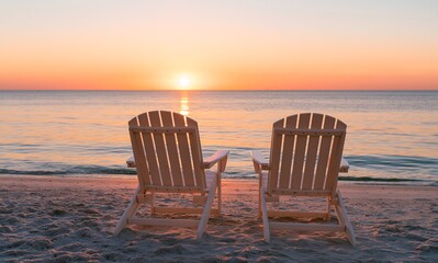 Fototapeta premium Two beach chairs seen from behind at sunset, with the sun on the horizon