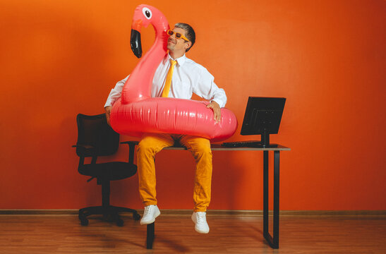 A smiling man sits on an office desk with a pink inflatable flamingo