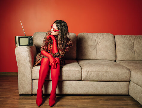 Stylish woman in leopard print dress and red gloves sitting on sofa with retro television