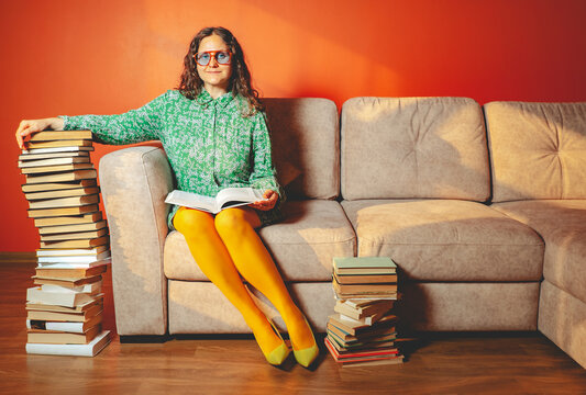 Woman in yellow tights reading on sofa with stacks of books indoors