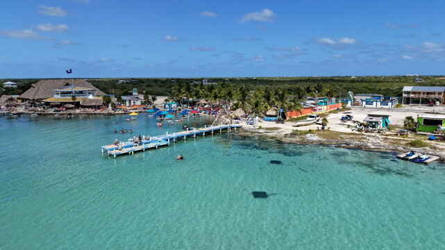 Aerial view of a wooden pier extending into the turquoise Caribbean Sea at San Pedro, with palm trees, beach umbrellas, and coastal buildings in San Pedro, Belize.