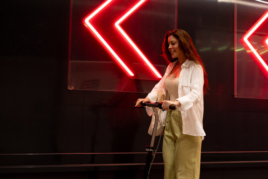Smiling woman riding electric scooter past neon lights in city