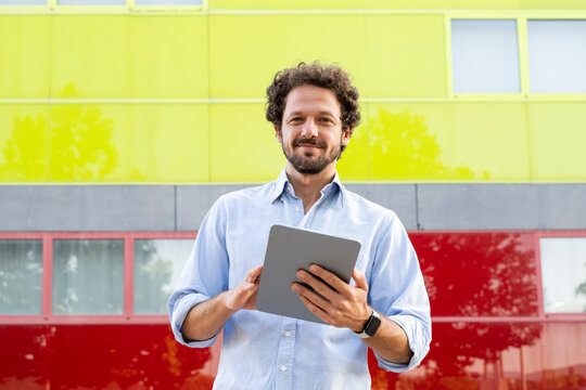 Male businessman with curly hair uses tablet by colored wall