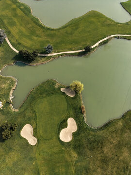 Aerial view of a golf course featuring green fairways, sand bunkers, and water hazards surrounded by trees and walking paths in Opera, Lombardy, Italy.