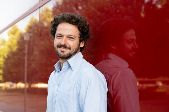 Smiling businessman in a blue shirt stands by a red wall