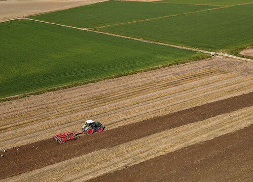 Aerial view of a tractor harrowing farmland with brown soil and green fields in Sesto Ulteriano, Lombardy, Italy.
