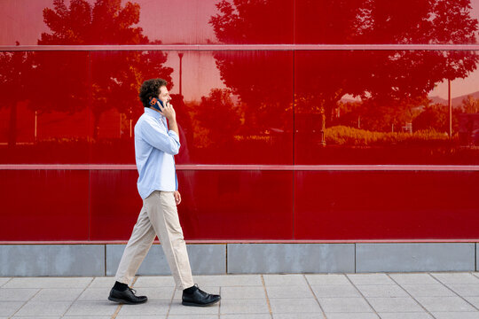 A businessman walks outdoors while using a smartphone near a red wall