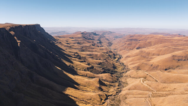 Aerial view of the rugged Sani Pass winding through the dramatic, golden-hued mountains of the Drakensberg range under a clear blue sky in Sani Pass, Lesotho.