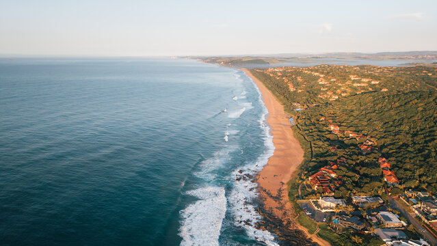 Aerial view of the coastline with sandy beaches, crashing ocean waves, lush coastal vegetation, and residential properties bathed in golden hour light in Ballito, KwaZulu-Natal, South Africa.
