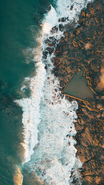 Aerial view of the rocky coastline with crashing waves and a natural tidal pool at sunset in Ballito, KwaZulu-Natal, South Africa.