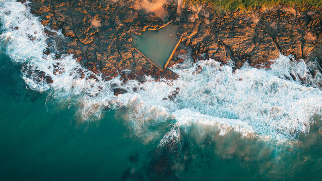 Aerial view of a natural tidal pool nestled in rocky coastline with crashing waves at golden hour in Ballito, KwaZulu-Natal, South Africa.