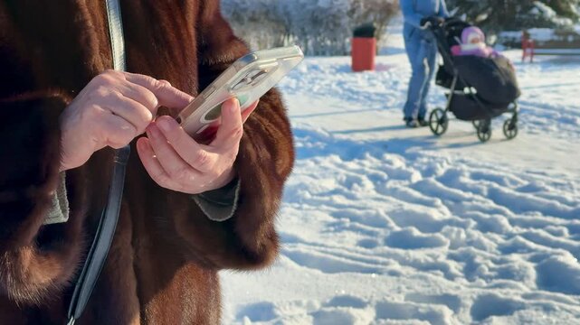 Woman in mink coat texting closeup, hands holding phone in snowy park, severe frost on fur, crossbody bag at side