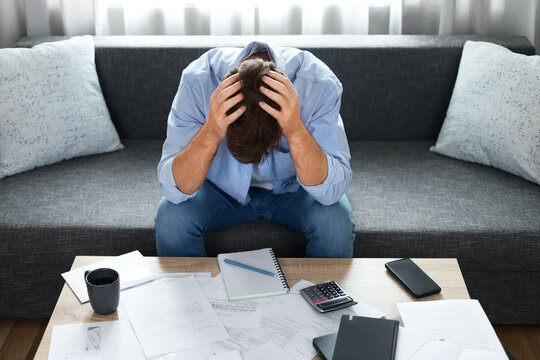 Stressed man holding his head in despair over unpaid utility bills. He faces severe financial debt, rising expenses, and an inflation crisis while sitting at home.