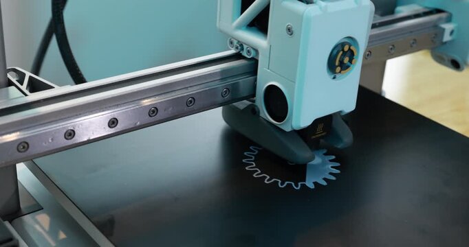 Close-up of a 3d printer printing a gear shape on a black build plate with a metal frame and light blue parts
