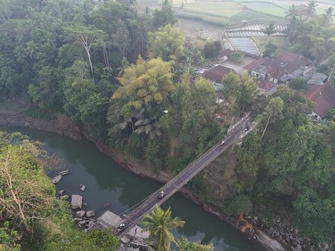 Aerial view of a suspension bridge spanning a river surrounded by lush tropical forest and rural buildings in Yogyakarta, Special Region of Yogyakarta, Indonesia.